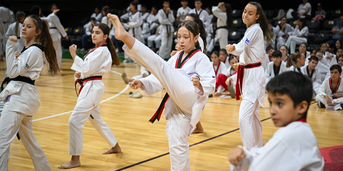 Kapalı bir spor salonunda tekvando kıyafetleri giymiş çocuklar ve gençler, siyah kuşak sınavı veya antrenmanı için bir arada bulunuyor. Ön planda, beyaz tekvando üniformalı ve siyah kuşaklı bir kız çocuğu havaya tekme atarken, yanındaki diğer öğrenciler de benzer kıyafetlerle duruş sergiliyor. Arka planda ise yerde oturan veya ayakta bekleyen çok sayıda sporcu ve izleyici yer alıyor.