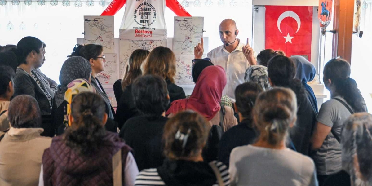 Beyaz gömlekli bir konuşmacı, bir grup kadına ve çocuğa hitap ediyor. Konuşmacının arkasında üzerinde 