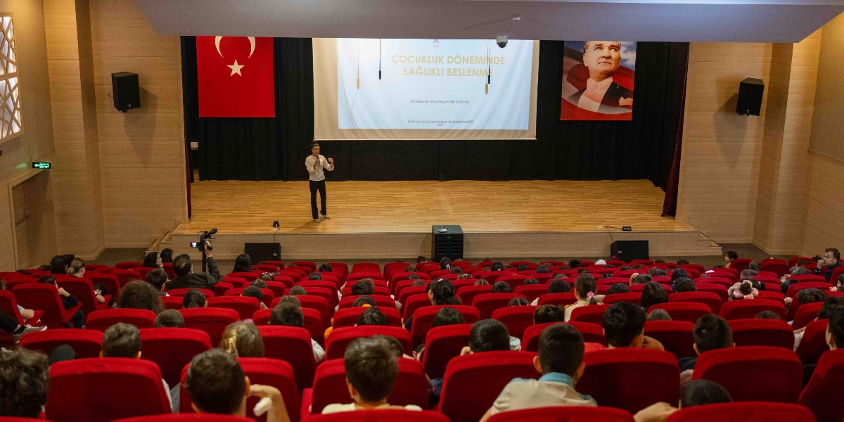 Modern bir konferans salonunda, sahnenin ortasında ayakta duran bir konuşmacı, kalabalık bir izleyici grubuna sunum yapıyor. Sahnenin arka planındaki dev ekranda 