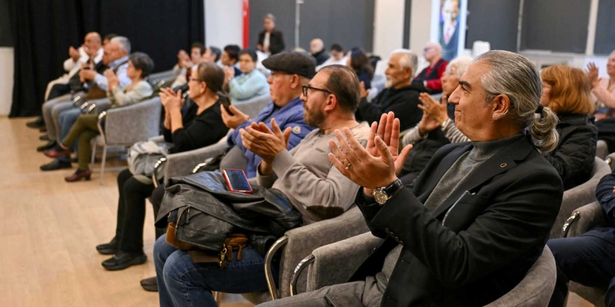 Konferans salonunda oturan ve alkışlayan izleyici kitlesi. Ön planda, arkadan toplanmış uzun saçlı, gri balıkçı yaka kazak ve ceket giyen bir adamın yan profili görülüyor. İzleyiciler arasında farklı yaş gruplarından kadın ve erkekler yer alıyor; bazıları ellerinde telefon tutuyor, bazıları ise dikkatle sahneye bakarak alkışlıyor. Salonun arka planında siyah perdeler ve aydınlık bir ortam bulunuyor.