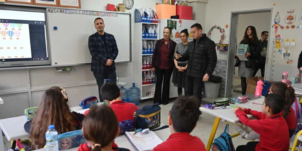 Bir ilkokul sınıfında, akıllı tahtanın önünde duran ekose ceketli bir adam öğrencilere hitap ediyor. Sınıfın içinde iki kadın öğretmen ve elinde bir kutu tutan başka bir kadın daha bulunuyor. Kırmızı okul forması giymiş öğrenciler sıralarında oturarak konuşmayı dinliyor. Sınıfın duvarlarında eğitici görseller ve raflarda okul malzemeleri yer alıyor.