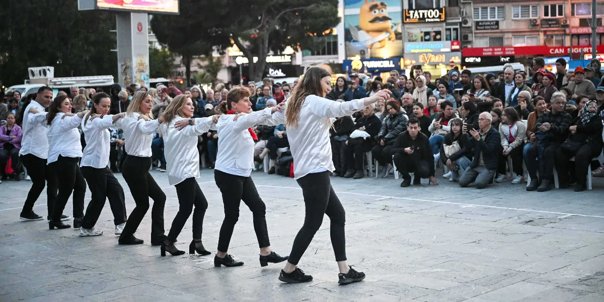 Bornova'da bir meydanda, beyaz gömlek ve siyah pantolon giymiş bir grup dansçı, birbirlerinin omuzlarına tutunarak oluşturdukları bir sıra halinde halay çekiyor. Dansçıların arkasında, onları izleyen kalabalık bir seyirci grubu oturuyor ve ayakta duruyor. Arka planda ise çeşitli dükkan tabelalarının bulunduğu bir şehir merkezi manzarası yer alıyor.