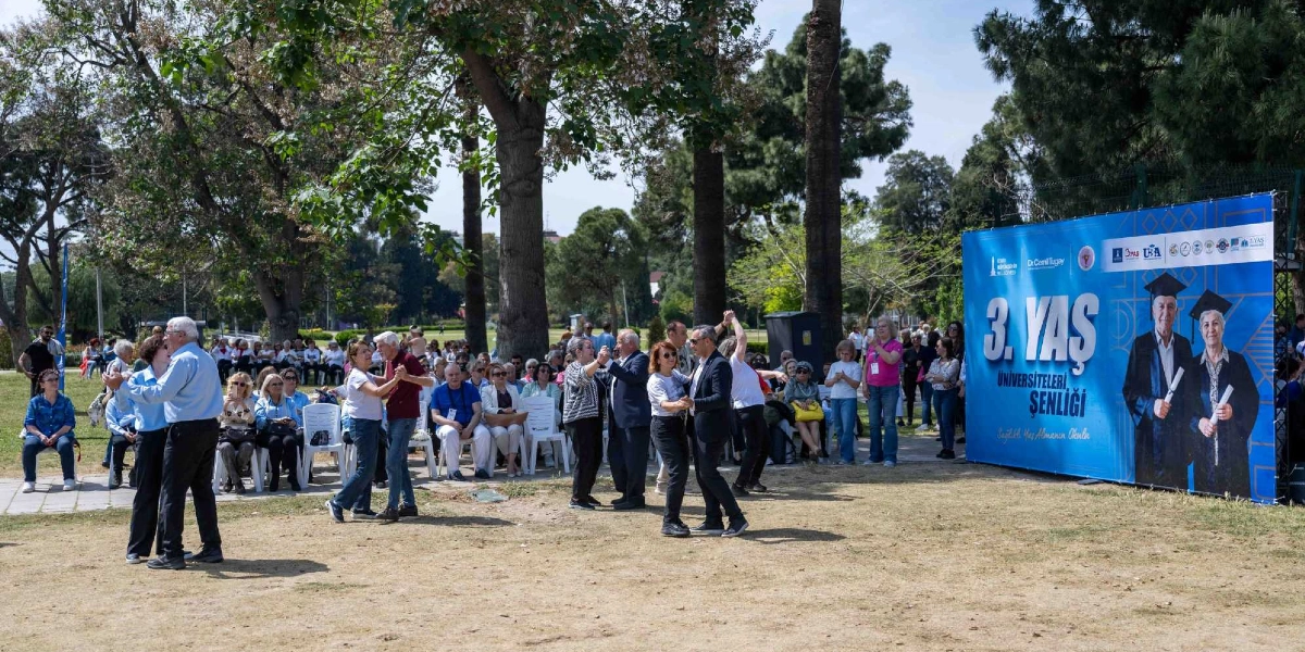 Açık havada, ağaçlık bir alanda düzenlenen 3. Yaş Üniversiteleri Şenliği'nde, mezuniyet cübbesi giymiş yaşlı bir kadın ve erkeğin fotoğrafının yer aldığı büyük bir afişin önünde dans eden yaşlı çiftler ve onları izleyen katılımcılar bulunuyor.