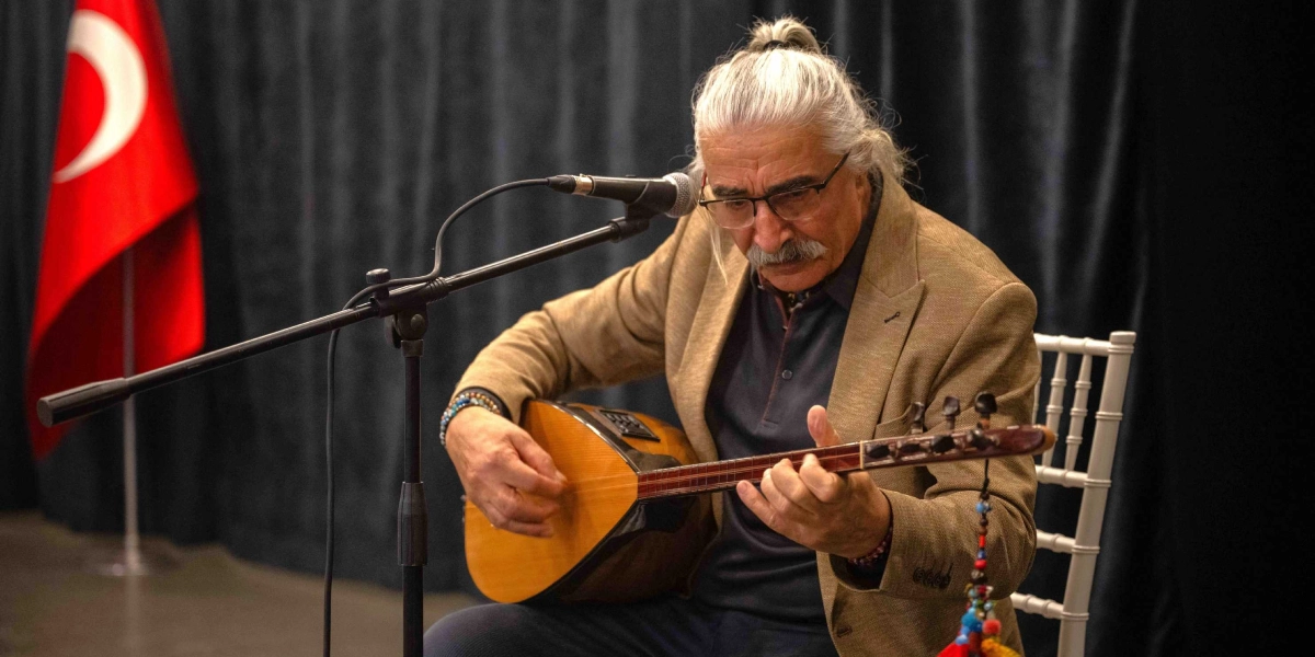 Gri saçlarını arkadan toplamış, gözlüklü ve bıyıklı bir müzisyen, koyu renkli bir sahne perdesinin önünde oturarak bağlama çalıyor. Müzisyen bej rengi bir ceket giyiyor ve önündeki mikrofona doğru hafifçe eğilmiş durumda. Arka planda, sol tarafta Türk bayrağı yer alıyor.