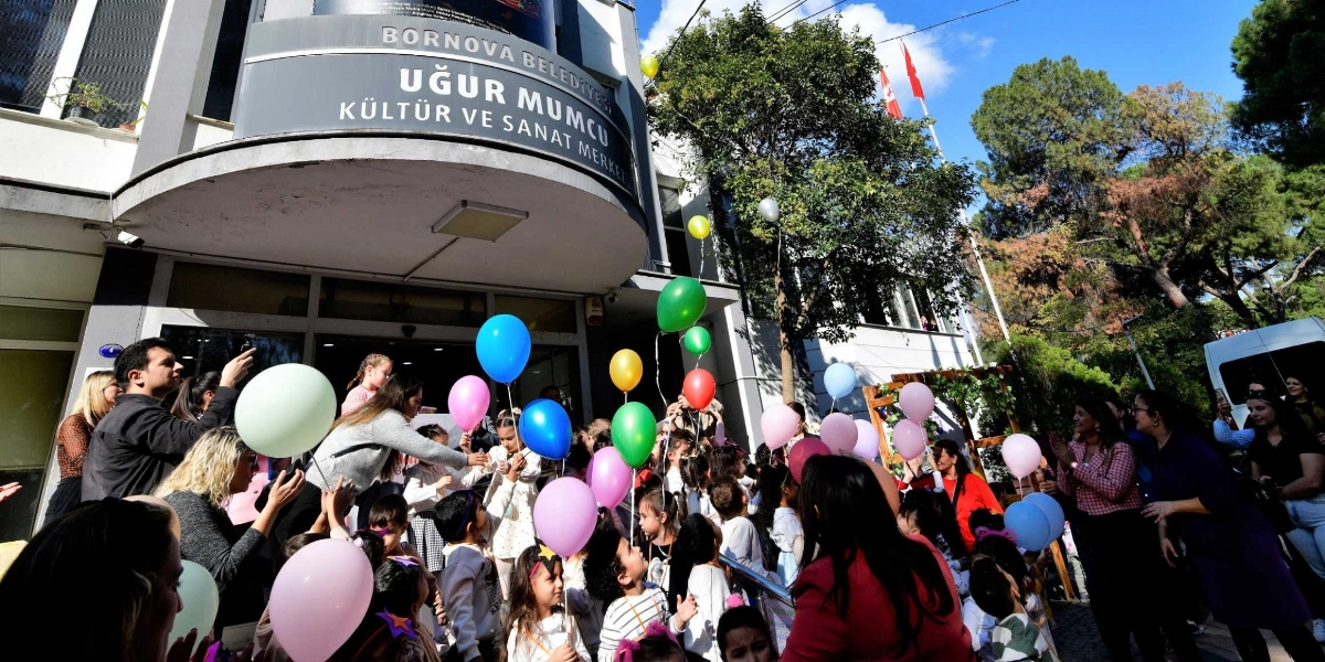 Bornova Belediyesi Uğur Mumcu Kültür ve Sanat Merkezi önünde toplanan çocuklar ve yetişkinler, ellerinde tuttukları rengarenk balonlarla Çocuk Hakları Günü'nü kutluyor. Binanın girişinde bir araya gelen grup, neşeli bir atmosferde balonları havaya kaldırıyor.
