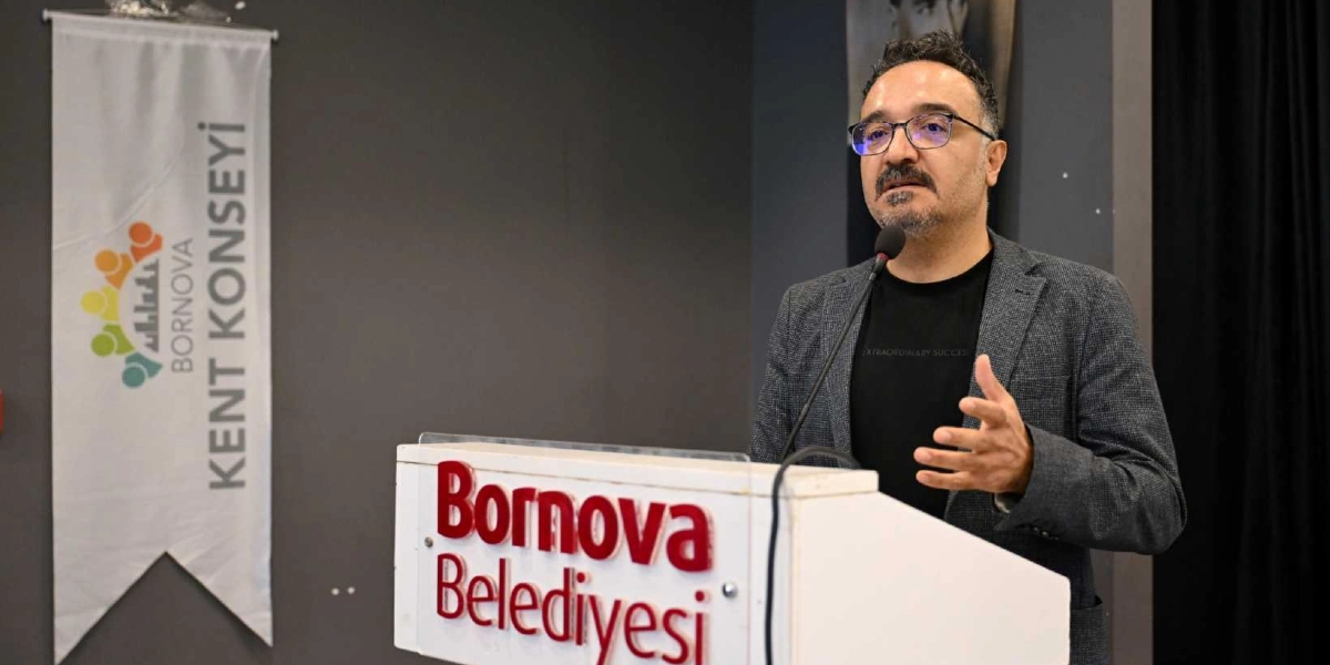 Gözlüklü, siyah tişört ve gri ceket giyen bir erkek, üzerinde Bornova Belediyesi yazan bir kürsüde konuşma yapıyor. Arka planda Bornova Kent Konseyi logosunun bulunduğu beyaz bir flama asılı duruyor.
