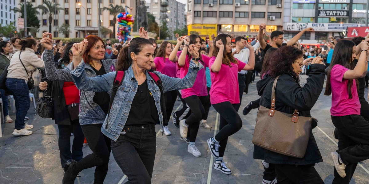 Bornova'da bir meydanda, Dünya Dans Günü kapsamında bir araya gelen farklı yaşlardan insanlar, kollarını havaya kaldırıp tek ayak üzerinde durarak senkronize bir şekilde dans ediyor. Katılımcıların birçoğu pembe tişörtler giyerken, ön planda kot ceketli bir kadın ve yanında omuzunda çantasıyla dans eden başka bir kadın dikkat çekiyor. Arka planda şehir binaları, dükkan tabelaları ve dansı izleyen kalabalık bir grup yer alıyor.