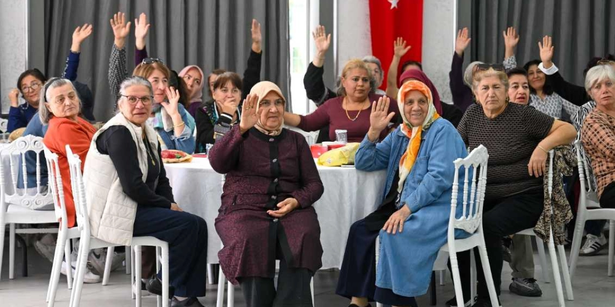Kapalı bir salonda beyaz örtülü masaların etrafında oturan, farklı yaş ve giyim tarzlarına sahip çok sayıda kadın, bir oylama veya onaylama amacıyla aynı anda ellerini havaya kaldırmış durumda. Kadınların bir kısmı başörtülü, bir kısmı ise günlük kıyafetler içinde; arka planda ise Türk bayrağı ve gri perdeler görülüyor.