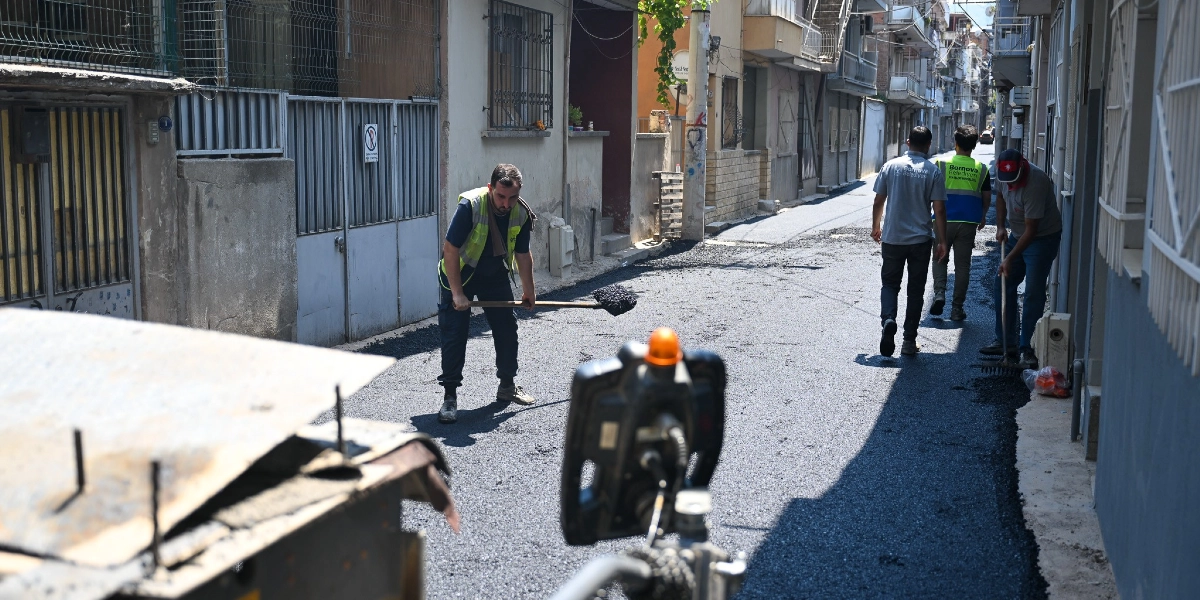 Bornova Belediyesi ekipleri, Çamdibi'ndeki bir sokakta asfalt serim ve yol yenileme çalışması yapıyor. İşçilerden biri elindeki kürekle asfaltı düzeltirken, diğerleri yolun kenarında çalışmaya devam ediyor.