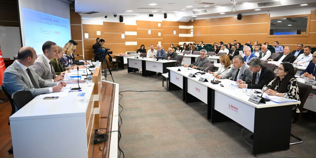 Bornova Belediyesi'nin Mayıs ayı olağan meclis toplantısında, U-şeklinde dizilmiş masalarda oturan meclis üyeleri ve yöneticiler yer alıyor. Salonun ön tarafında, üzerinde Bornova Belediyesi logosu bulunan beyaz masalarda oturan yetkililer toplantıyı yönetirken, arka sıralarda diğer meclis üyeleri oturuyor. Sol taraftaki projeksiyon perdesinde 