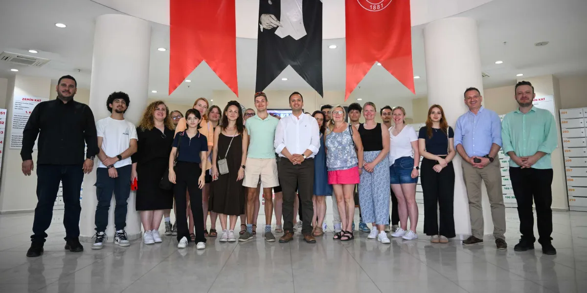 Aydınlık bir iç mekanda, yan yana duran farklı yaş ve milletlerden oluşan kalabalık bir gençlik grubu, arkalarında asılı olan Atatürk silüetli siyah bir bayrak ve iki yanında kırmızı bayrakların önünde poz veriyor. Grup üyeleri günlük kıyafetler içinde gülümseyerek kameraya bakıyor; arka planda ise beyaz sütunlar ve üzerinde numaralar bulunan posta kutusu benzeri bölmeler yer alıyor.