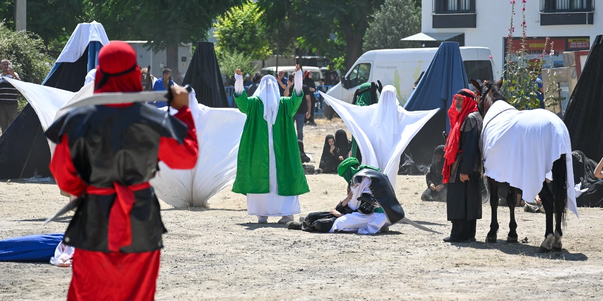 Açık havada düzenlenen Kerbela anma töreninde, tarihi kıyafetler giymiş oyuncular Kerbela olayını canlandırıyor. Ön planda kırmızı kıyafetli ve kılıçlı bir figür, orta alanda ellerini havaya kaldırmış yeşil cübbeli bir kişi ve yerde oturan başka figürler bulunuyor. Sağ tarafta ise beyaz örtüyle kaplanmış bir at ve yanında geleneksel kıyafetli bir kişi yer alıyor. Arka planda siyah çadırlar, izleyiciler ve beyaz bir minibüs görülüyor.