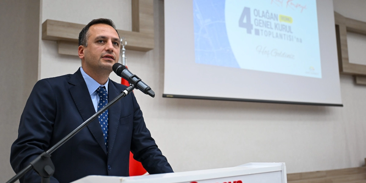 Koyu renk takım elbiseli ve mavi kravatlı bir erkek, kürsüde mikrofon başında konuşma yapıyor. Arka plandaki projeksiyon perdesinde 
