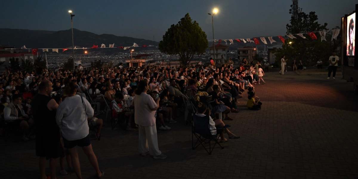 Akşam saatlerinde açık havada kurulan bir sinema ekranının önünde toplanmış kalabalık bir grup insan, sandalyelerde oturarak veya ayakta durarak izleme yapıyor. Arka planda şehir ışıkları ve dağ silüeti görülürken, alanın üzerinde Türk bayrakları ve flamalar asılı. Ekranın sağ tarafında bir animasyon karakterinin görüntüsü yansıtılıyor.
