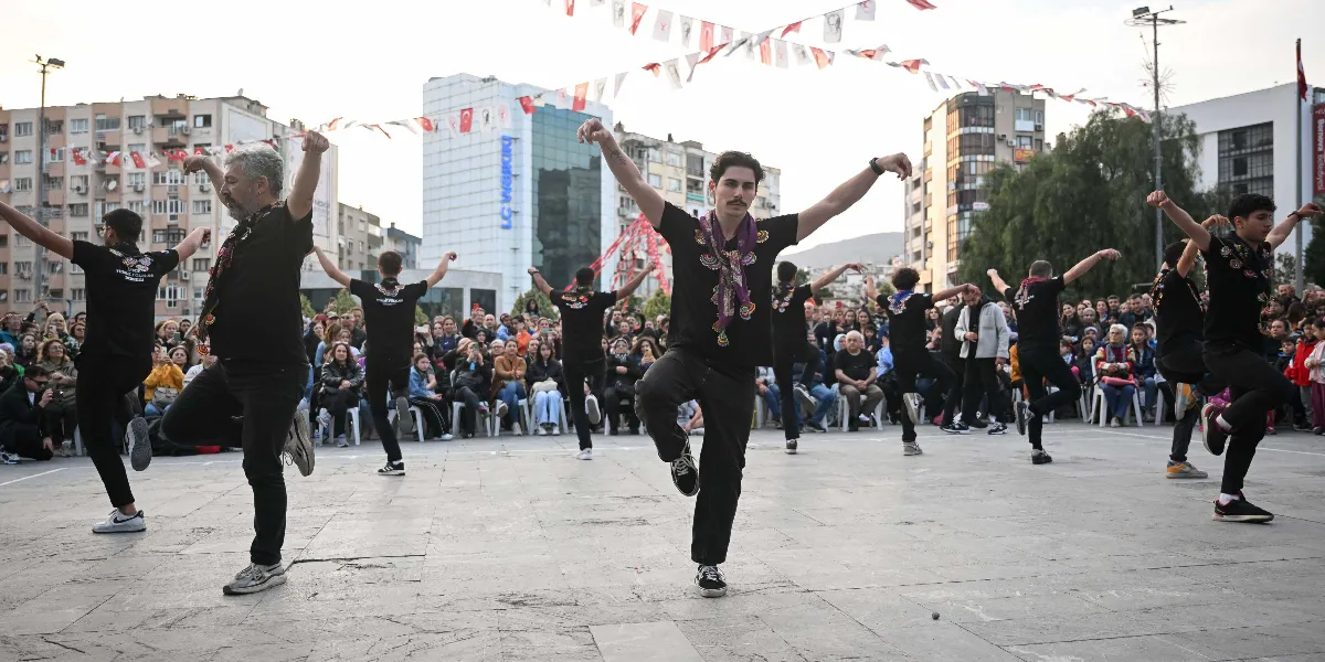 Bornova'da bir meydanda, siyah tişört ve siyah pantolon giymiş bir grup erkek dansçı, geleneksel bir halk oyununu sergiliyor. Dansçılar kollarını iki yana açmış, tek ayak üzerinde durarak figürlerini icra ediyor. Arka planda onları izleyen kalabalık bir seyirci grubu bulunuyor ve meydan, üzerinde Türk bayrakları asılı iplerle süslenmiş durumda.