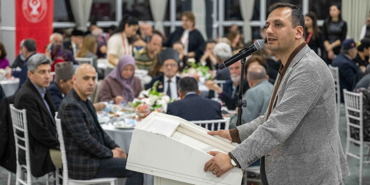 Kapalı bir mekanda düzenlenen yemekli bir etkinlikte, kürsüde konuşma yapan bir erkek. Konuşmacının önünde mikrofon bulunuyor ve arka planda masalarda oturan kalabalık bir davetli grubu yer alıyor. Masalarda oturanlar arasında başörtülü kadınlar, takım elbiseli erkekler ve çeşitli yaş gruplarından insanlar bulunuyor. Arka planda Bornova Belediyesi'ne ait olduğu anlaşılan kırmızı renkli bir flama göze çarpıyor.