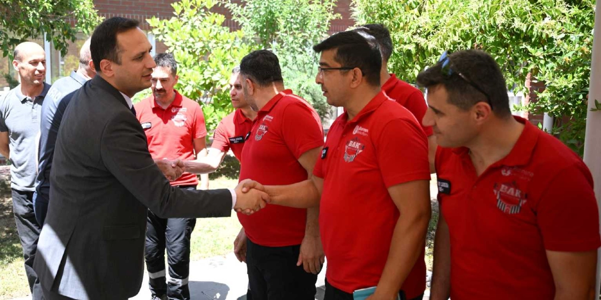Takım elbiseli bir yetkili, üzerinde Bornova Belediyesi BAK (Bornova Arama Kurtarma) logolu kırmızı tişörtler bulunan bir grup arama kurtarma personeliyle açık havada tokalaşıyor.