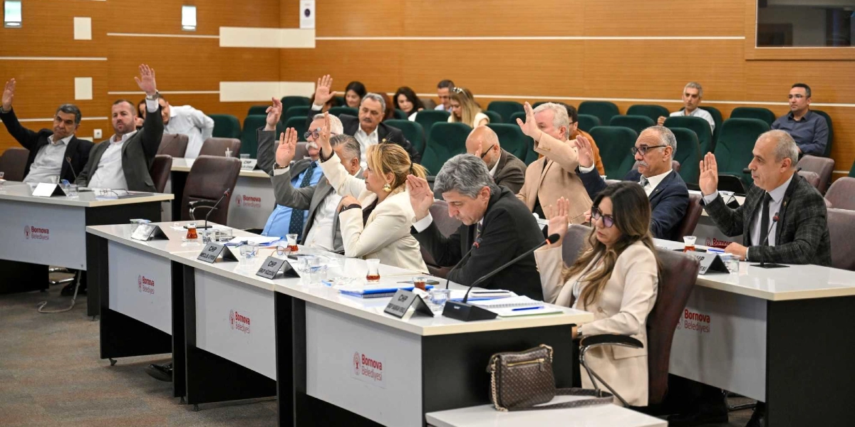 Bornova Belediyesi meclis salonunda, sıralarda oturan çok sayıda meclis üyesi ellerini kaldırarak oylama yapıyor. Üyeler, önlerinde Bornova Belediyesi logolu masaların bulunduğu, ahşap panellerle kaplı modern bir toplantı salonunda yer alıyor. Masaların üzerinde isimlikler, mikrofonlar ve çay bardakları bulunuyor.