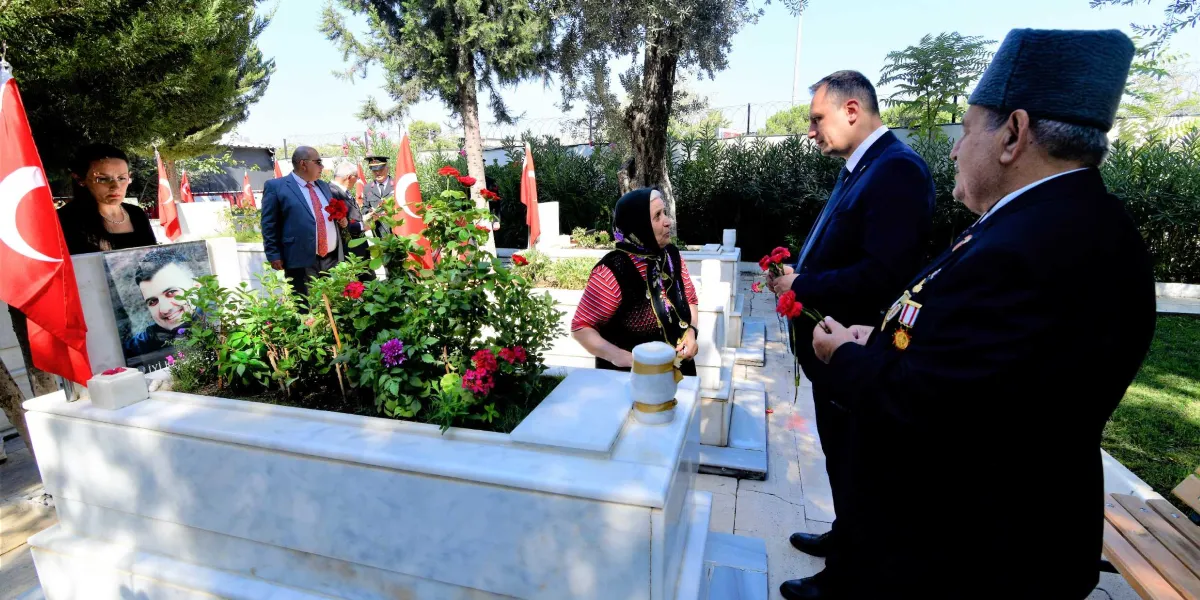 Açık havada, mermer bir şehitlikte, üzerinde genç bir erkeğin fotoğrafı bulunan mezar başında toplanmış bir grup insan. Başörtülü yaşlı bir kadın, takım elbiseli bir adam ve göğsünde madalyaları bulunan kalpaklı bir gazi, ellerinde kırmızı karanfillerle mezarı ziyaret ediyor. Arka planda Türk bayrakları ve ağaçlar görülüyor.