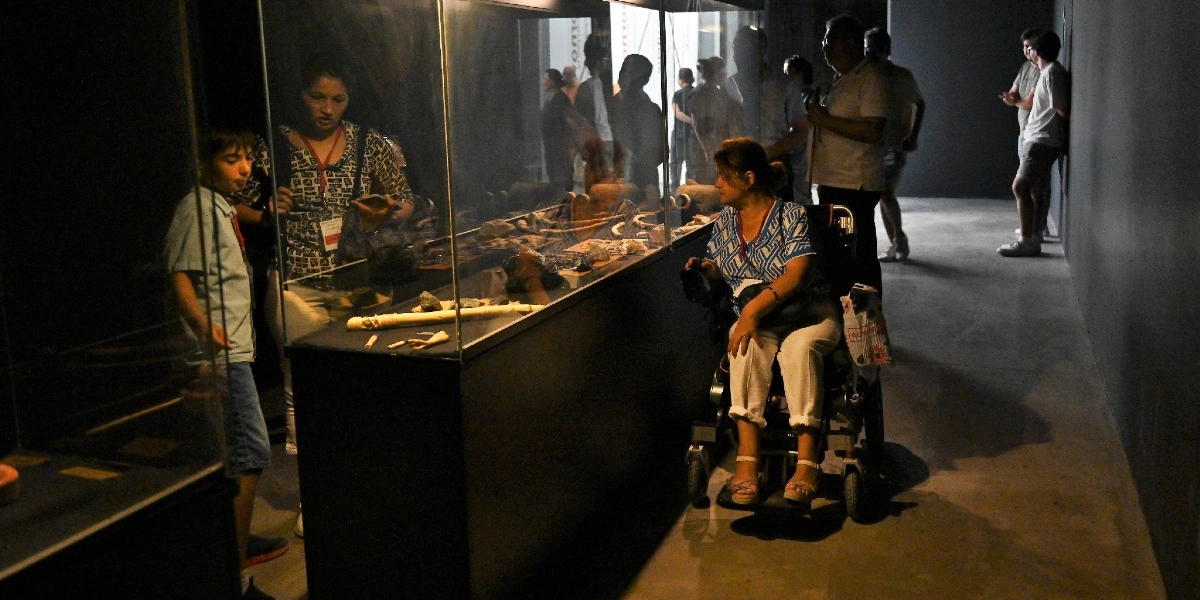 Müze ortamında, cam bir vitrin içerisinde sergilenen arkeolojik eserleri inceleyen ziyaretçiler. Tekerlekli sandalyedeki bir kadın, yanında duran bir çocuk ve diğer ziyaretçiler vitrindeki tarihi objelere bakıyor. Loş ışıklandırılmış sergi salonunda insanlar vitrinin etrafında toplanmış durumda.