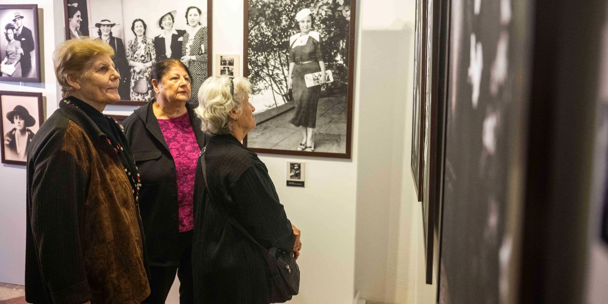 Şapka Devrimi'nin 100. yılına özel düzenlenen sergiyi gezen üç kadın, duvarlarda asılı olan ve dönemin şapkalı kadınlarını gösteren siyah beyaz fotoğrafları inceliyor. Kadınlar, sergi salonunda yan yana durarak çerçevelenmiş tarihi fotoğraflara dikkatle bakıyor.