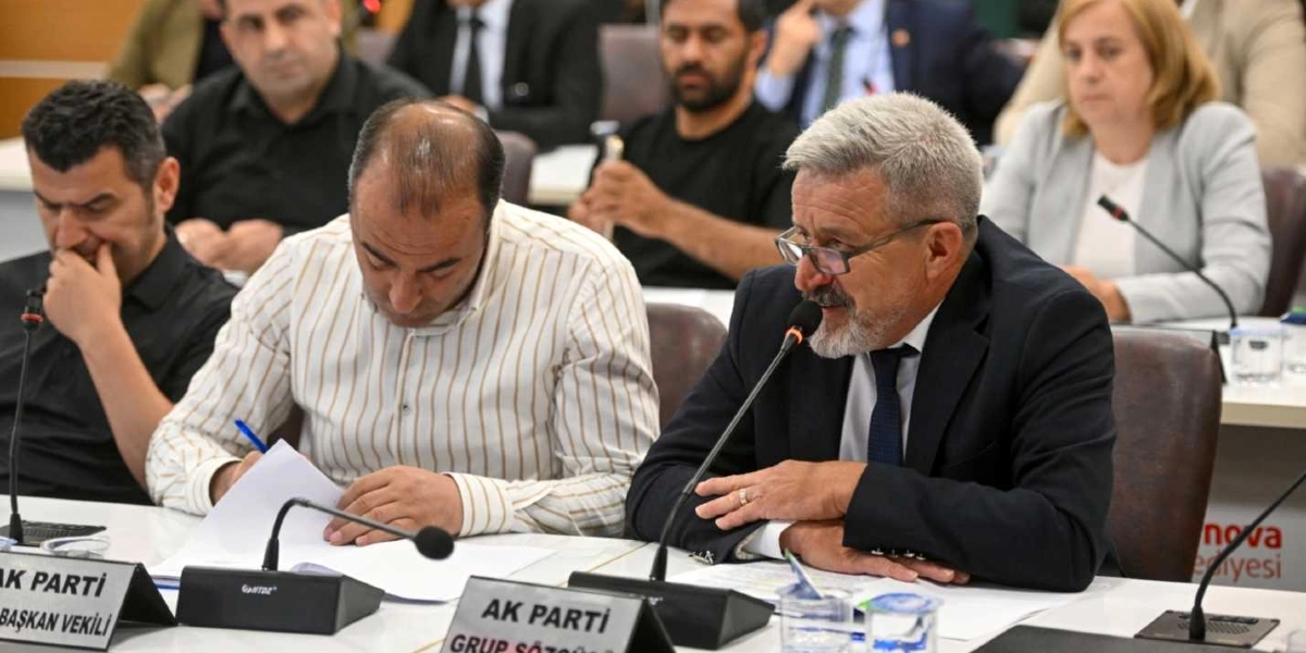 Resmi bir toplantı masasında oturan, önünde mikrofon bulunan ve konuşma yapan gri saçlı, gözlüklü bir erkek ile yanında not alan çizgili gömlekli bir kişi ve diğer katılımcılar. Masanın üzerinde AK Parti Grup Sözcüsü ve Başkan Vekili yazılı isimlikler yer alıyor.