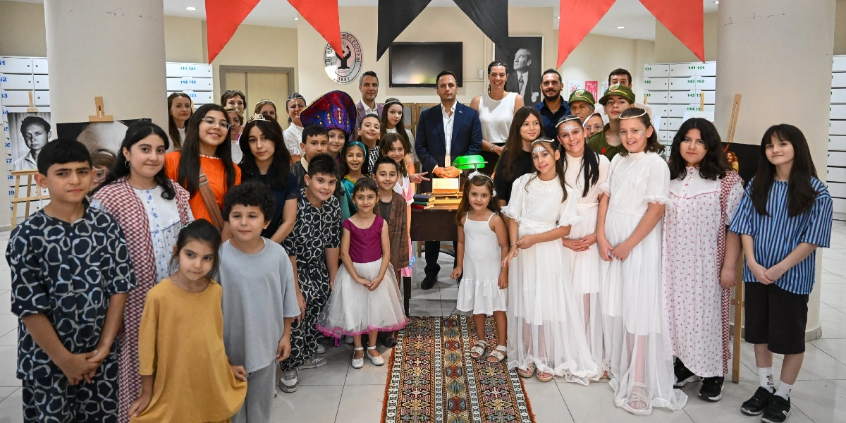 Bornova Belediyesi binasında, çeşitli yaş gruplarından çocukların ve yetişkinlerin bir arada bulunduğu bir sanat etkinliği. Çocukların bir kısmı beyaz elbiseler, taçlar ve özel kostümler giymiş; bazıları ise günlük kıyafetleriyle yer alıyor. Arka planda Bornova Belediyesi logosu, Atatürk portresi ve üzerinde kitaplar bulunan bir çalışma masası görülüyor. Mekanın tavanında kırmızı ve siyah süslemeler asılı, yanlarda ise üzerinde siyah beyaz fotoğrafların olduğu şövaleler bulunuyor. Grup, bir sanat sergisi veya gösteri sonrası hatıra fotoğrafı çektirmek için bir araya gelmiş.