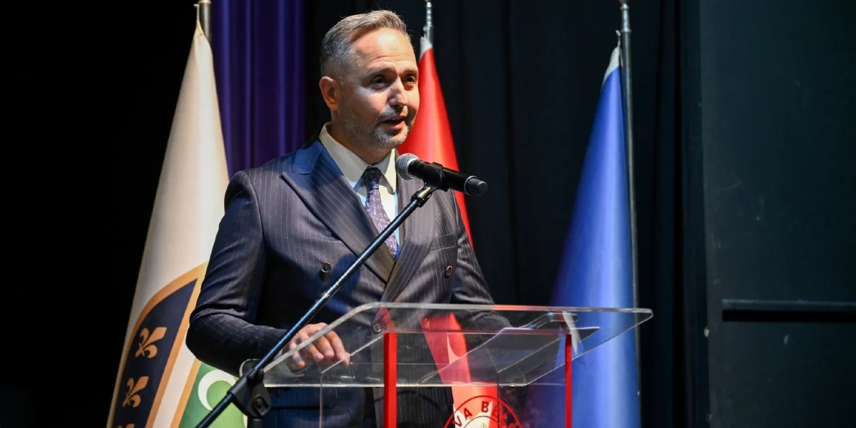 Koyu renkli çizgili takım elbise ve kravat giymiş, kısa saçlı ve hafif sakallı bir erkek, kürsüde mikrofon başında konuşma yapıyor. Arka planda Türk bayrağı ve üzerinde sarı zambak motifleri bulunan mavi-yeşil renkli bir bayrak yer alıyor.
