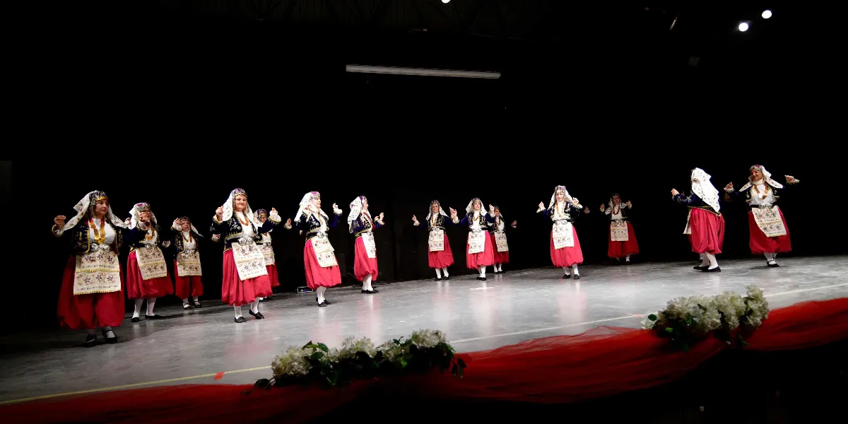 Siyah bir sahne üzerinde geleneksel halk oyunları kıyafetleri giymiş bir grup kadın dansçı, yan yana dizilmiş şekilde performans sergiliyor. Dansçılar, kırmızı etekler, işlemeli beyaz önlükler, koyu renkli yelekler ve başlarında beyaz örtülerle yer alıyor. Sahnenin ön kısmında beyaz çiçeklerden oluşan süslemeler ve kırmızı bir tül detayı bulunuyor.
