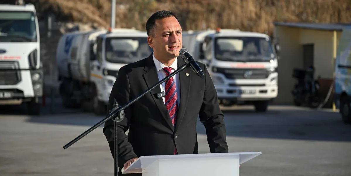 Siyah takım elbiseli ve kırmızı çizgili kravatlı bir erkek, açık havada, arkasında belediyeye ait hizmet araçlarının bulunduğu bir alanda kürsüde konuşma yapıyor.