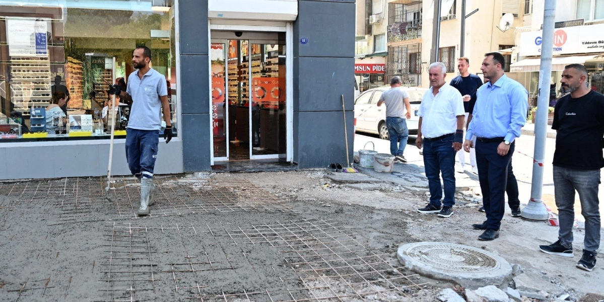 Bir optik mağazasının önünde devam eden kaldırım yenileme çalışmasında, gri tişörtlü ve çizmeli bir işçi elindeki aletle beton zemin üzerinde çalışıyor. Çalışma alanının hemen yanında, biri beyaz gömlekli, biri açık mavi gömlekli ve biri siyah tişörtlü olmak üzere üç erkek ayakta durarak çalışmaları izliyor. Arka planda ise başka bir işçinin yürüdüğü, dükkan vitrinlerinin ve cadde üzerindeki diğer iş yerlerinin göründüğü bir sokak manzarası yer alıyor.