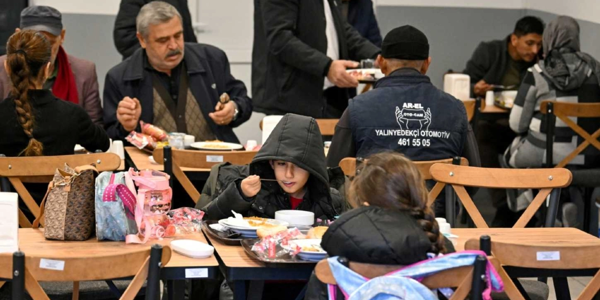 Kapalı bir mekanda, ahşap masalarda yemek yiyen bir grup insan. Ön planda kapüşonlu mont giymiş bir çocuk yemek yerken, yanında sırt çantası olan başka bir çocuk oturuyor. Hemen arkalarında lacivert yelekli bir adam ve diğer masalarda yemek yiyen yetişkinler bulunuyor. Yeleğin üzerinde 