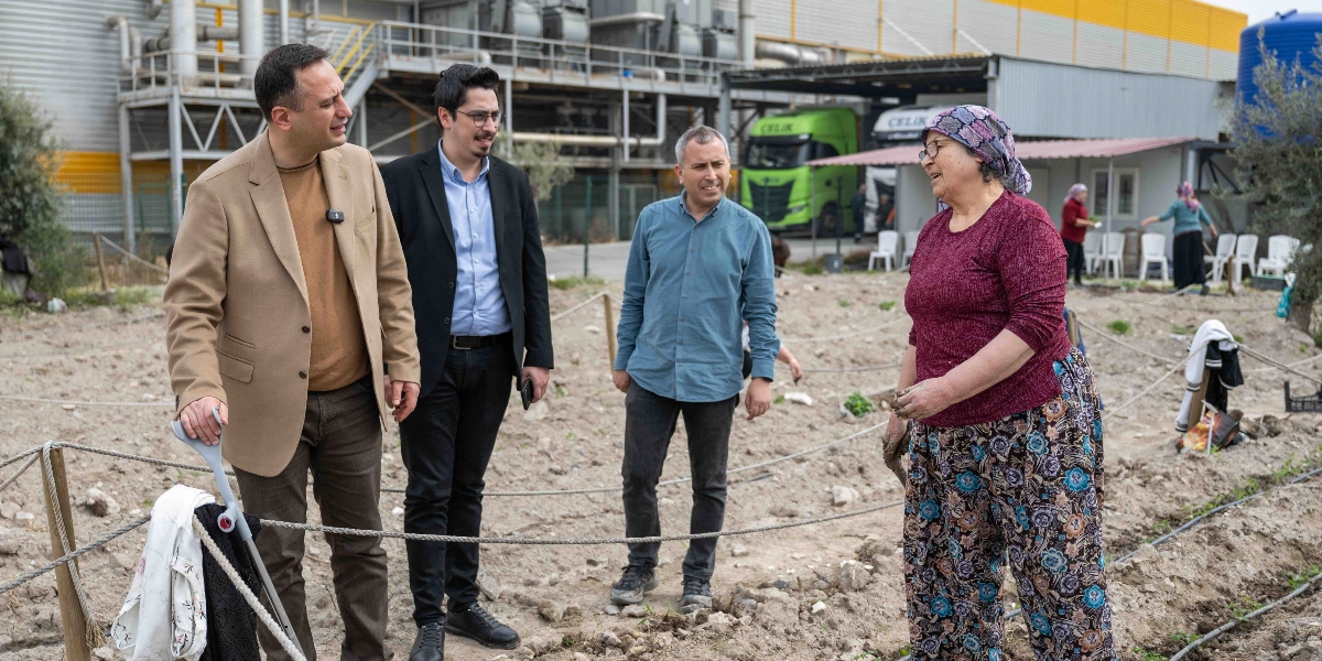 Açık havada, toprak bir alanda, elleri çamurlu olan başörtülü bir kadın, yanındaki üç erkekle sohbet ediyor. Erkeklerden biri bej ceketli, diğeri siyah ceketli, üçüncüsü ise mavi gömlekli. Arka planda büyük bir endüstriyel tesis, park halindeki tırlar ve başka kişilerin bulunduğu bir bahçe alanı görülüyor. Bej ceketli adamın yanında bir koltuk değneği bulunuyor.