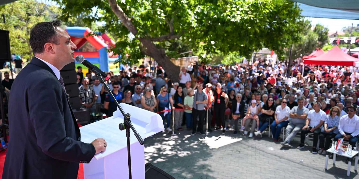 Açık havada, ağaçların gölgesinde düzenlenen bir etkinlikte, kürsüde konuşma yapan takım elbiseli bir erkek ve onu dinleyen kalabalık bir topluluk yer alıyor. Arka planda kırmızı çadırlar ve çocuklar için şişme oyun parkı görülüyor.
