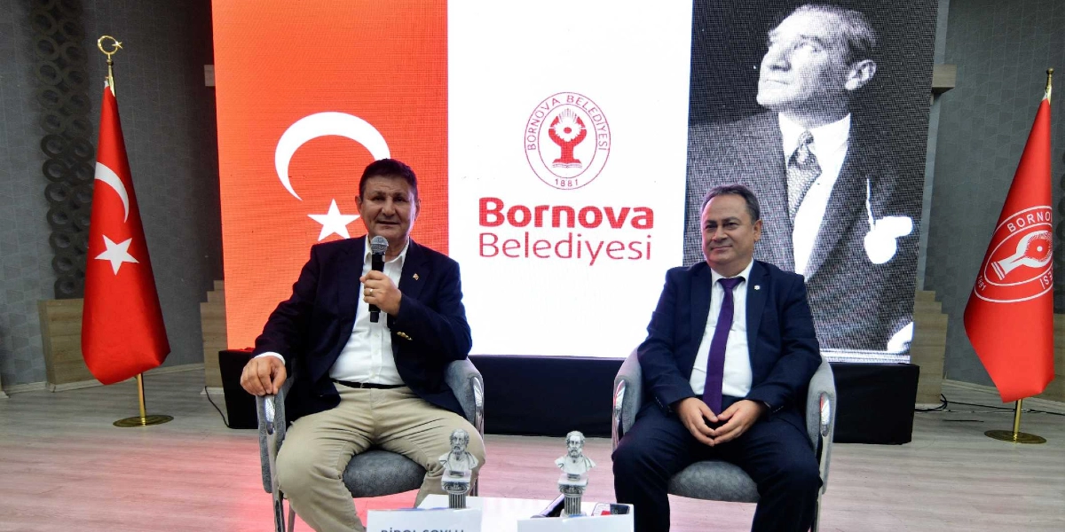 Kapalı bir salonda, arka planda dev bir ekranda Türk bayrağı, Bornova Belediyesi logosu ve Mustafa Kemal Atatürk'ün portresi yer alırken, iki erkek yan yana oturuyor. Soldaki kişi elinde mikrofon tutarak konuşuyor, sağdaki kişi ise onu dinliyor. Her iki katılımcının önünde küçük birer büst ve isimlik bulunuyor. Sahnenin sağ ve sol köşelerinde ise Türk bayrağı ve Bornova Belediyesi bayrağı yer alıyor.