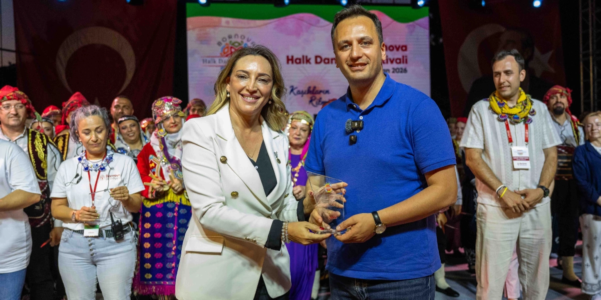 Bornova Halk Dansları Festivali sahnesinde, beyaz ceketli bir kadın, mavi polo yaka tişört giyen bir erkeğe plaket takdim ediyor. Arka planda geleneksel kıyafetler giymiş dansçılar ve Türk bayrakları yer alıyor. Sahne üzerindeki dijital ekranda 