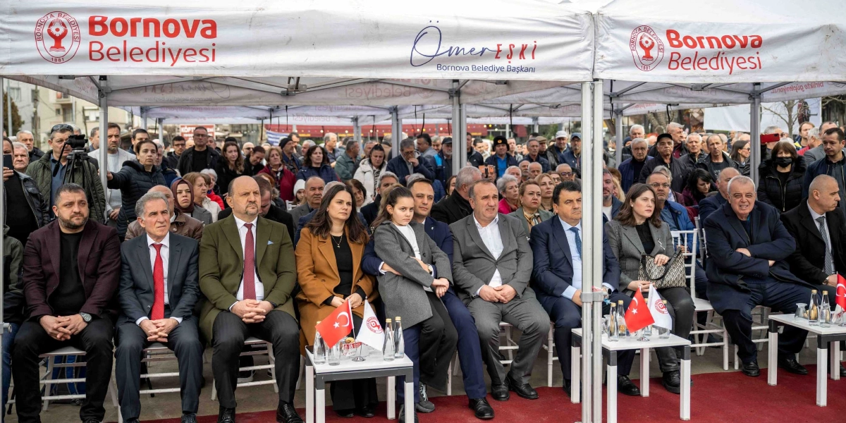 Bornova Belediyesi tarafından düzenlenen açık hava etkinliğinde, üzerinde Bornova Belediyesi logoları ve Ömer Eşki isminin yer aldığı beyaz çadırların altında oturan protokol üyeleri ve kalabalık bir izleyici grubu görülüyor. Ön sırada takım elbiseli erkekler ve şık giyimli kadınlar yan yana otururken, aralarında bir çocuk da bulunuyor. Masaların üzerinde küçük Türk bayrakları ve Bornova Belediyesi logolu flamalar yer alıyor. Arka planda ise etkinliği takip eden çok sayıda vatandaş bulunuyor.