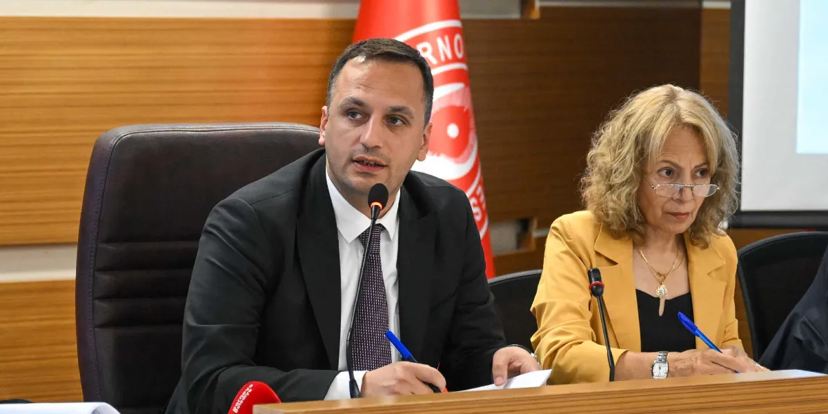 Bornova Belediye Başkanı Mustafa İduğ, bir toplantı masasında mikrofon önünde konuşma yaparken, yanında sarı ceketli bir kadın katılımcı not alıyor. Arka planda Bornova Belediyesi'nin kırmızı beyaz renkli logosunun bulunduğu bir bayrak yer alıyor.