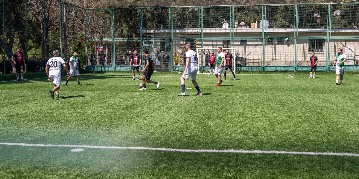 Açık havada, tel örgülerle çevrili suni çim sahada oynanan bir futbol maçında, farklı formalar giymiş oyuncular topun etrafında pozisyon alıyor. Beyaz formasıyla 16 numaralı oyuncu topu sürerken, siyah ve yeşil-beyaz formalı diğer oyuncular oyun alanına dağılmış durumda. Sahayı çevreleyen tellerin arkasında maçı izleyen bir grup seyirci bulunuyor.