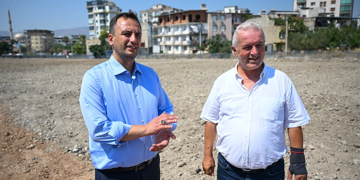 Açık renkli gömlek giymiş iki erkek, arkalarında şehir binalarının göründüğü boş ve topraklı bir arazide yan yana durarak gülümsüyor. Soldaki kişi mavi gömlekli, sağdaki kişi ise beyaz gömlekli ve elinde siyah bir bileklik takılı.