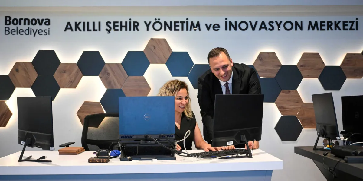 Bornova Belediyesi Akıllı Şehir Yönetim ve İnovasyon Merkezi'nde, beyaz bir çalışma masasının başında oturan bir kadın ve yanında ayakta duran bir erkek, bilgisayar ekranına bakarak gülümseyerek çalışıyor. Arka planda, üzerinde 