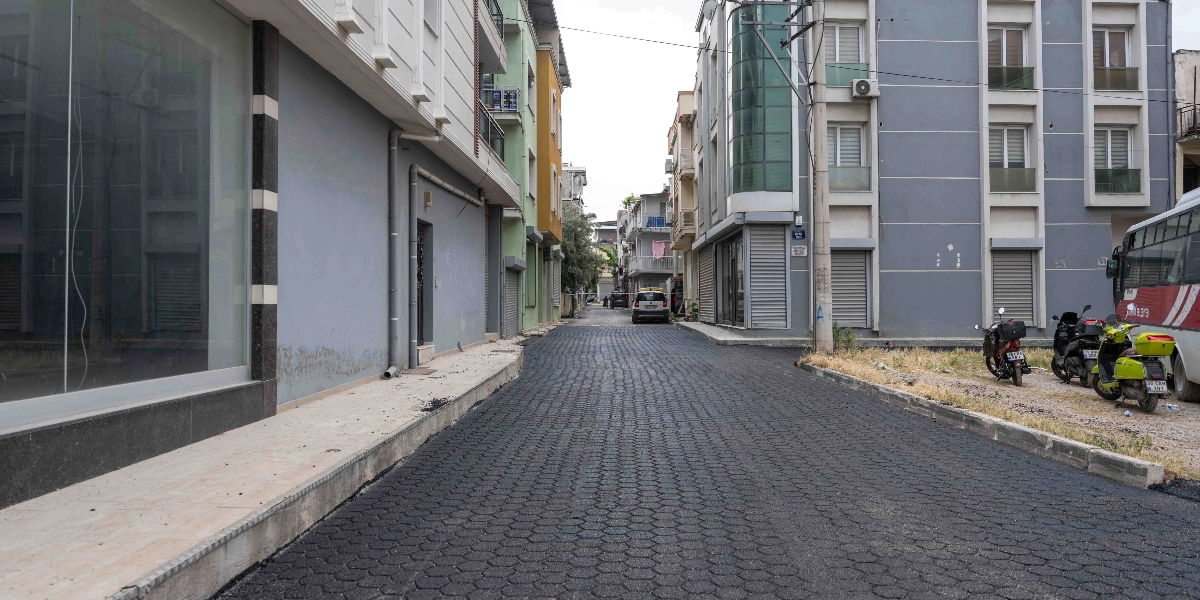 Bornova'da yeni asfaltlanmış, petek dokulu yüzeye sahip bir sokak. Sokağın iki yanında çok katlı apartmanlar, sağ tarafta park halindeki motosikletler ve bir otobüsün arka kısmı, ileride ise yolun ortasında duran bir otomobil görülüyor.