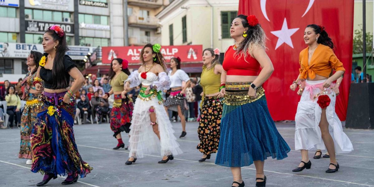 Bornova'da açık havada düzenlenen Dünya Dans Günü kutlamasında, geleneksel ve modern dans kostümleri giymiş bir grup kadın dansçı, arkalarında asılı büyük bir Türk bayrağı önünde performans sergiliyor. Dansçılar renkli etekler, bellerinde madeni para detaylı kemerler ve saçlarında çiçek aksesuarlarıyla dans figürlerini icra ederken, arka planda onları izleyen bir kalabalık ve şehir merkezi binaları görülüyor.