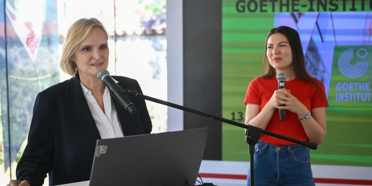 Kürsüde konuşma yapan sarı saçlı ve siyah ceketli bir kadın ile yanında duran, kırmızı tişörtlü ve uzun kahverengi saçlı genç bir kadın. Arka planda üzerinde Goethe-Institut logosu ve yazısı bulunan yeşil renkli bir ekran yer alıyor.