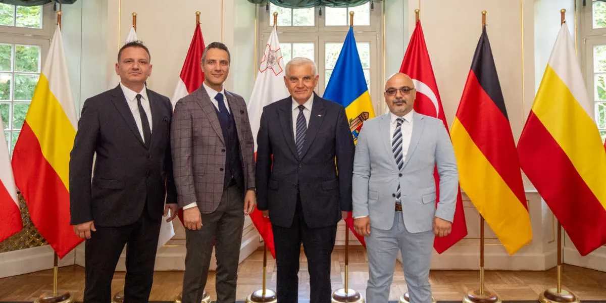 Farklı ülkelerin bayraklarının önünde yan yana duran dört erkek temsilci. Soldan sağa doğru; koyu renk takım elbiseli, gri ekose ceketli, lacivert takım elbiseli ve açık gri takım elbiseli dört kişi, resmi bir toplantı ortamında objektife gülümseyerek bakıyor. Arka planda Malta, Moldova, Türkiye ve Almanya gibi çeşitli ülkelerin bayrakları sıralanmış durumda.