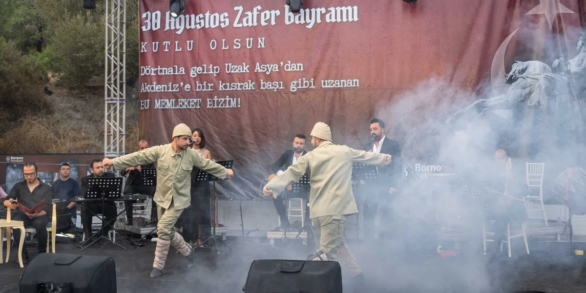 30 Ağustos Zafer Bayramı kutlamaları kapsamında sahnede, Kurtuluş Savaşı dönemi asker kıyafetleri giymiş iki kişi halk oyunu sergiliyor. Sahnenin arka planında üzerinde 