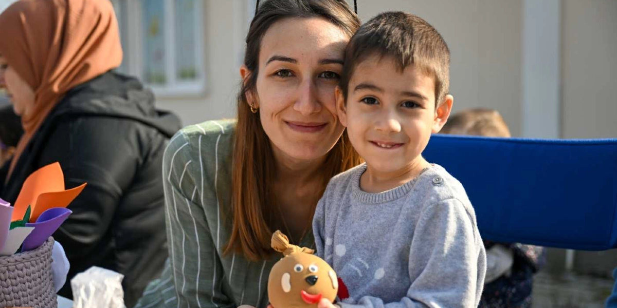 Yeşil çizgili bir bluz giyen genç bir kadın ve gri kazaklı küçük bir erkek çocuk, ellerinde tuttukları kendi yaptıkları çim adamla birlikte kameraya gülümseyerek poz veriyor. Arka planda ise başörtülü bir kadın ve el işi malzemeleriyle dolu bir sepet görülüyor.