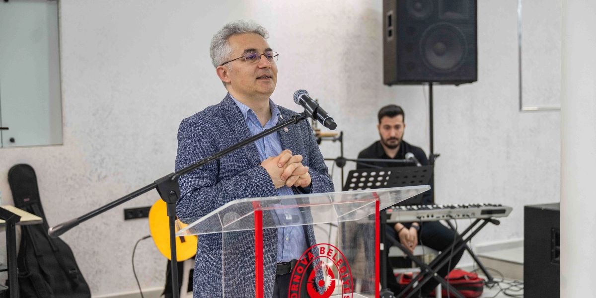 Gözlüklü, gri saçlı bir erkek, üzerinde Bornova Belediyesi logosu bulunan şeffaf bir kürsüde mikrofon başında konuşma yapıyor. Arka planda ise siyah kıyafetli bir müzisyen klavye başında oturuyor ve yanında bir gitar kılıfı ile hoparlör sistemi bulunuyor.