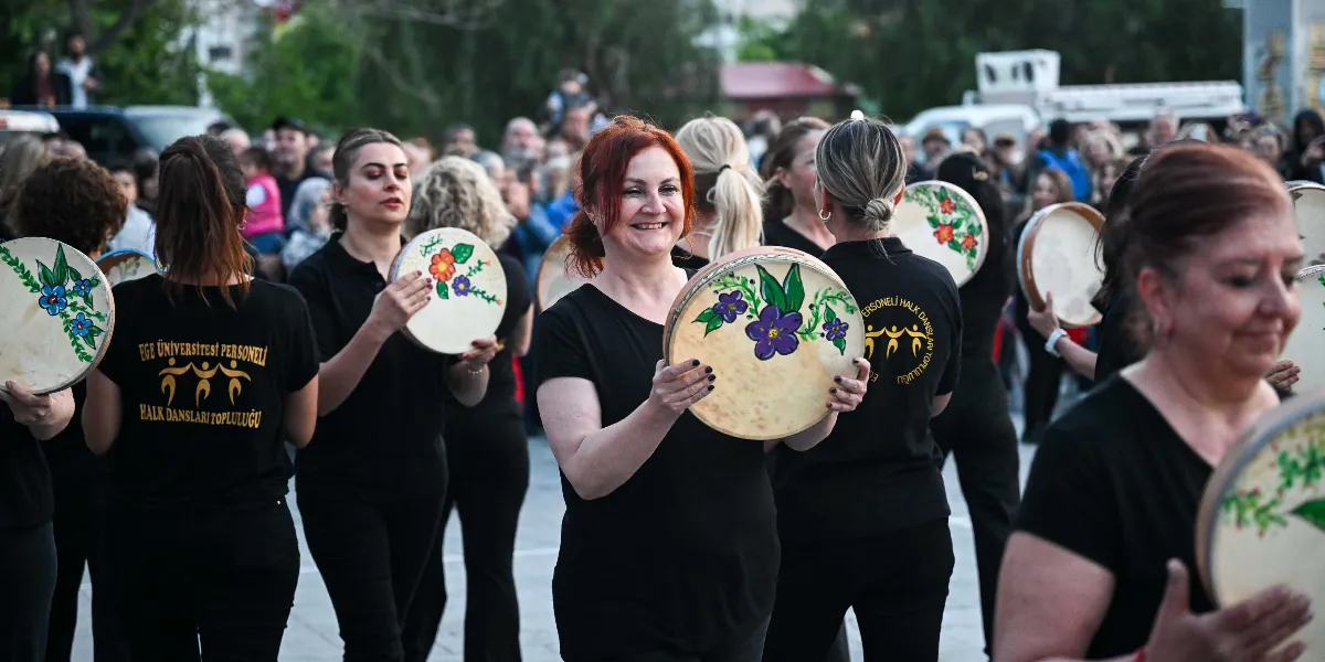 Ege Üniversitesi Personeli Halk Dansları Topluluğu üyeleri, üzerinde çiçek desenleri bulunan teflerle açık havada gösteri yapıyor. Siyah tişörtlü dansçılar, ellerinde tuttukları teflerle gülümseyerek yürürken, arkalarında kalabalık bir izleyici grubu yer alıyor.