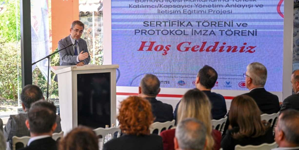 Gri ceketli bir konuşmacı, kürsüde katılımcılara hitap ediyor. Arkasındaki büyük ekranda 